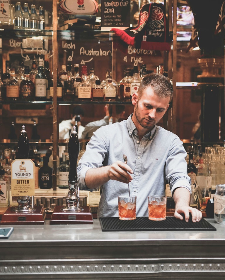 A bartender is preparing two cocktails behind a well-stocked bar, featuring a variety of liquor bottles.