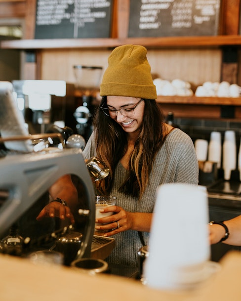 A young woman wearing a yellow beanie and glasses is smiling while preparing a drink at a coffee shop.